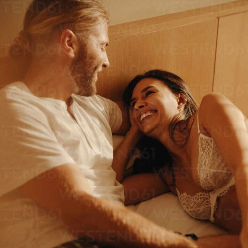 Couple in a candid and intimate moment, lying and laughing together on a bed. The warm, soft lighting adds to the cozy and romantic atmosphere. This photo has intentional use of 35mm film grain.